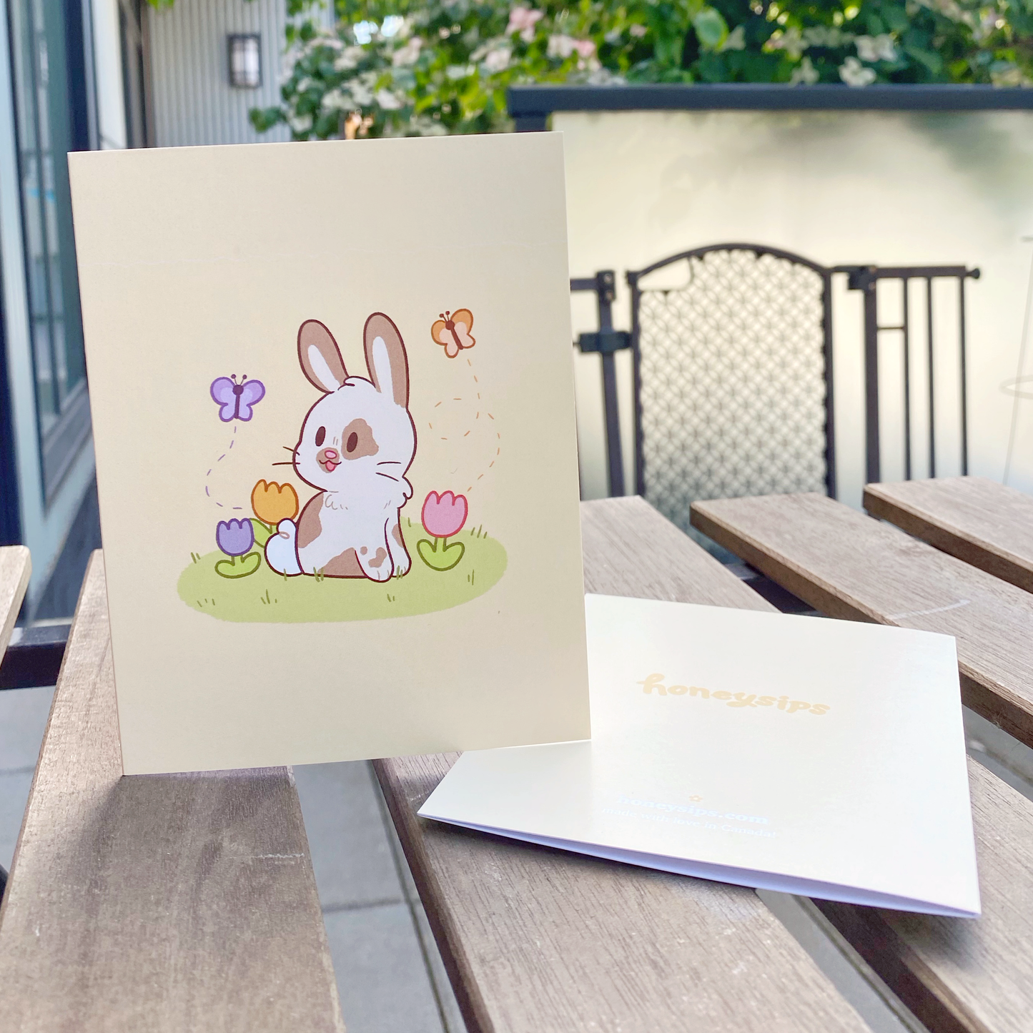A2 pastel yellow greeting card with a white and brown spotted bunny with 3 purple, orange, and pink tulips, and a purple and orange butterfly. Photographed on a brown wooden slatted table