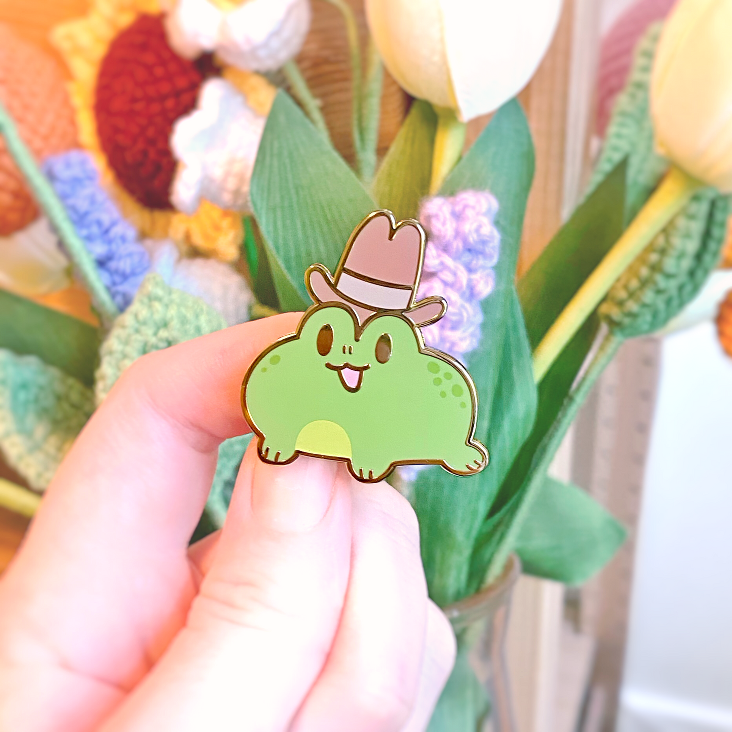 Gold plated enamel pin of a bight green frog smiling with an open mouth wearing a tall brown cowboy hat. Photographed against fake green plants and pastel crocheted flowers.