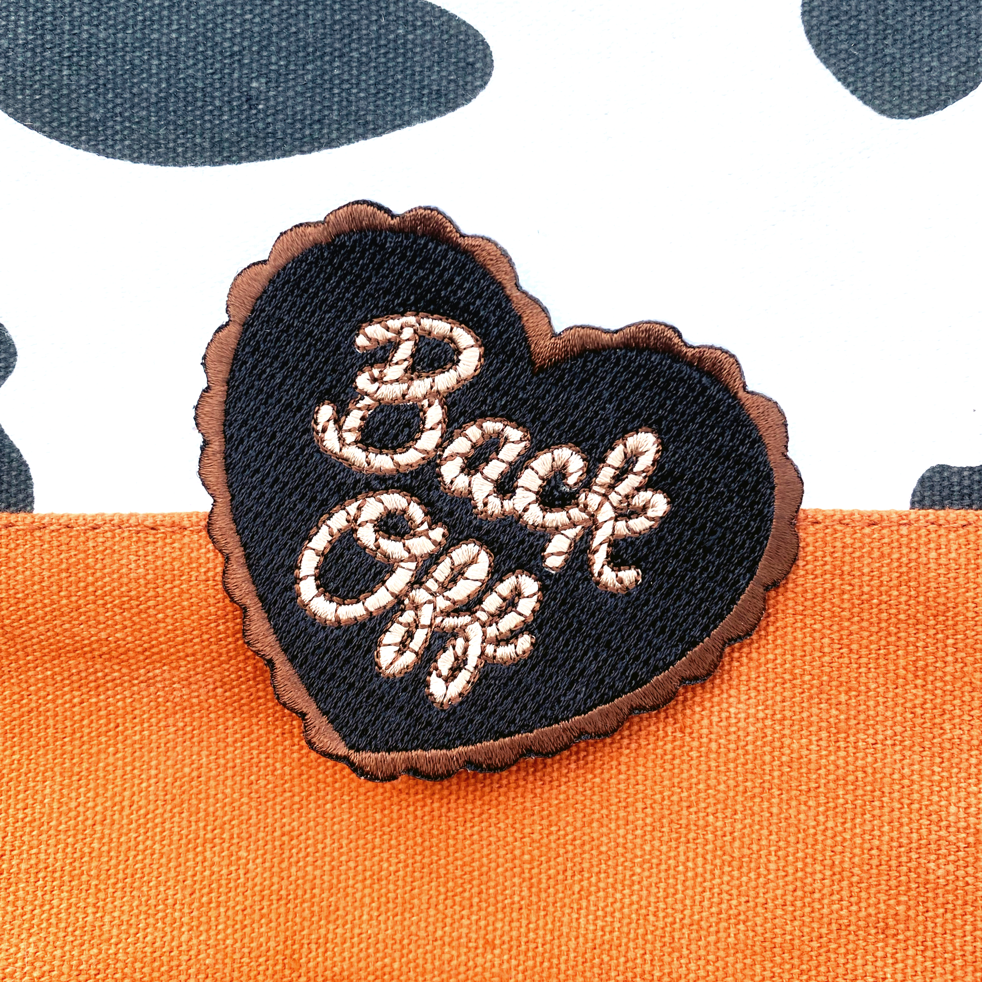 Black and yellow heart shaped embroidered patch with the quote "Back Off" in cursive photographed against a black and white cow print background