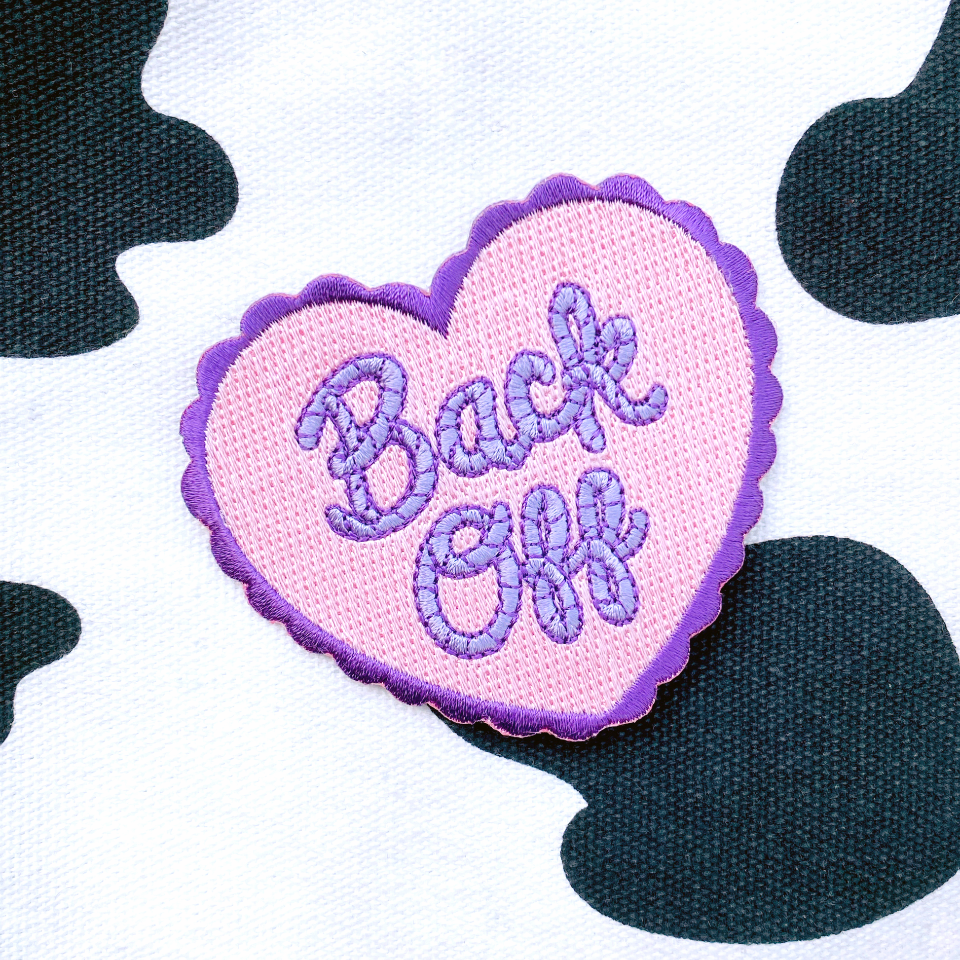 Pink and purple heart shaped embroidered patch with the quote "Back Off" in cursive photographed against a black and white cow print background