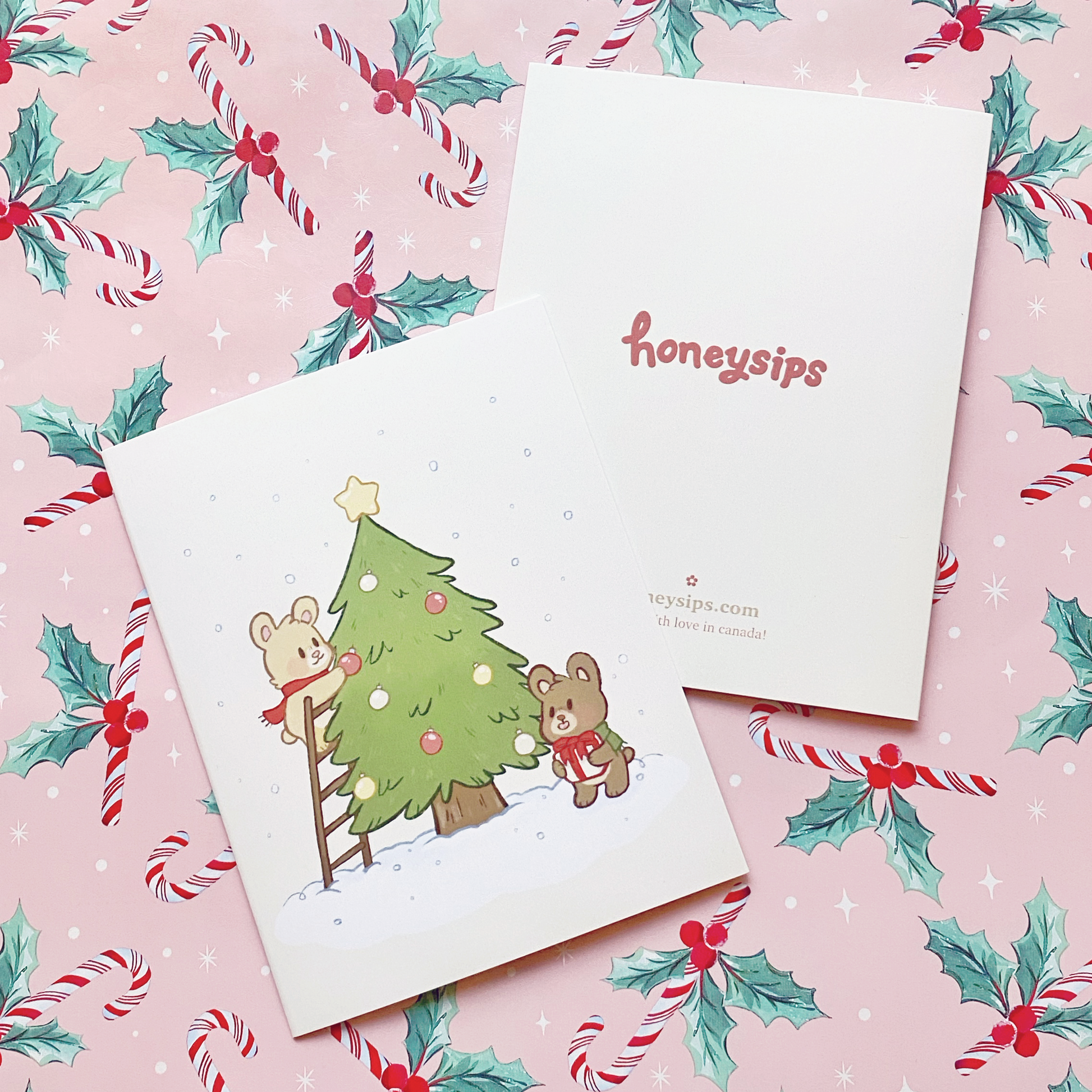 A2 Christmas card with two brown bears decorating a green Christmas tree in the snow on a cream card. Photographed on pastel pink wrapping paper with red and white candy canes on it with bunches of green and red holly.