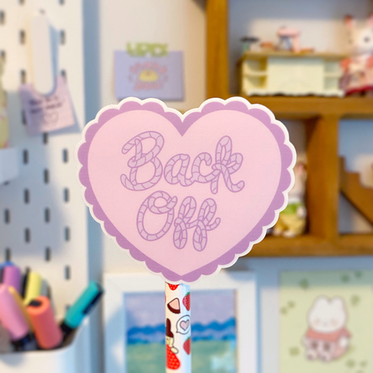 Pink and purple heart-shaped vinyl sticker with the quote "Back Off" in cursive on it, photographed in front of a colourful cluttered background