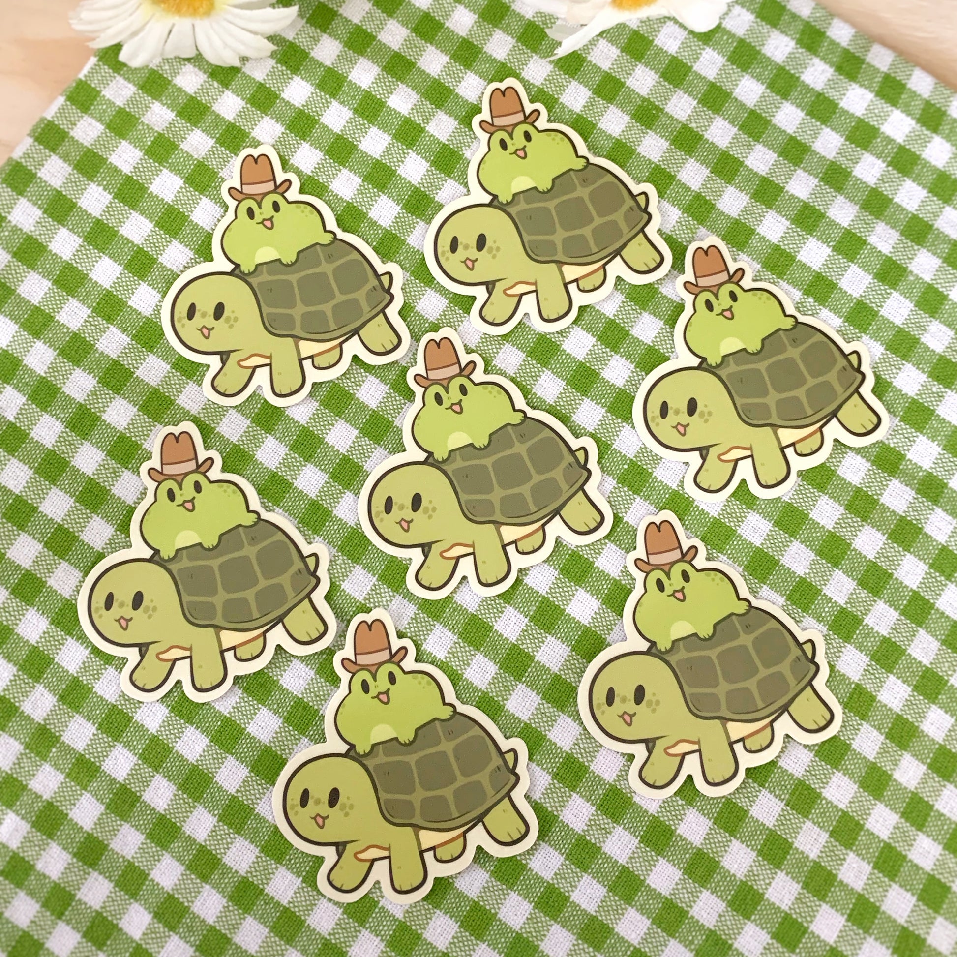 7 vinyl stickers of an illustrated green frog with an open mouth smile wearing a tall brow cowboy hat, riding a green turtle with an open smile. Photographed against a green and white gingham cloth.