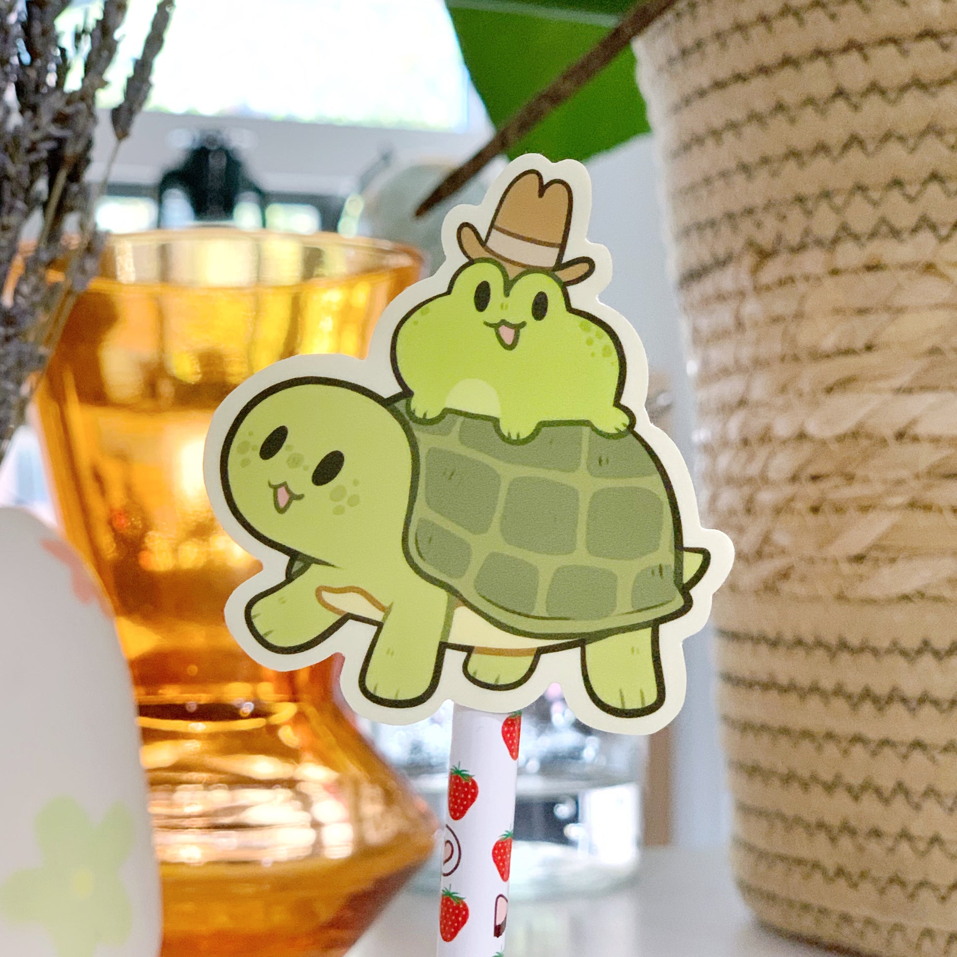 Vinyl sticker of an illustrated green frog with an open mouth smile wearing a tall brow cowboy hat, riding a green turtle with an open smile. Photographed in front of an orange transparent vase, and a large wicker basket.