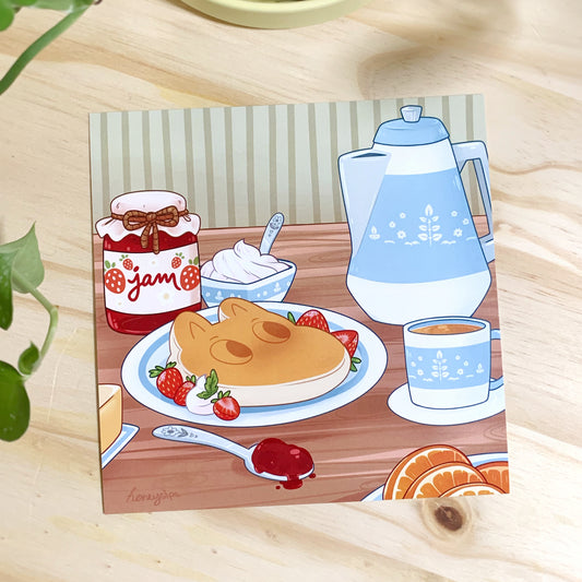 Printed illustration of a breakfast scene featuring a red strawberry jar of jam, cup of whipped cream, hippo shaped pancake with strawberries, spoon with jam, vintage blue and white coffee carafe, vintage blue and white coffee cup, and some orange slices on a wooden table with green pinstriped wallpaper. Photographed on a wooden background with a house plant to the left for decor.