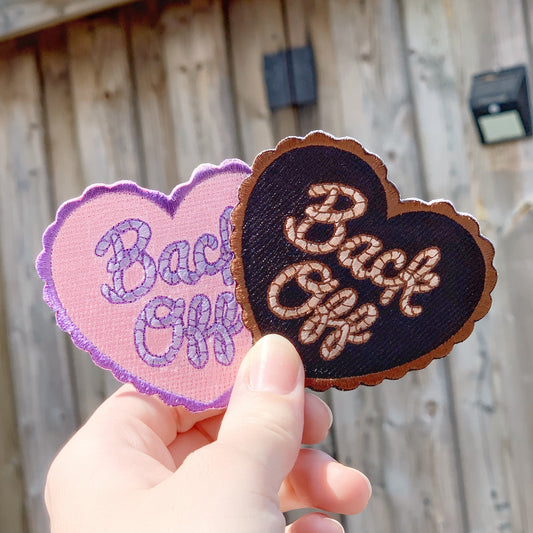Two heart-shaped embroidered patches (one pink and purple, and one black and yellow), with the quote "Back Off" in cursive held up in front of a wooden shed