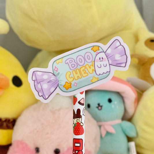 Pastel purple, teal, and yellow vinyl sticker shaped as a wrapped candy with a cute smiling ghost and the quote "Boo Chew" on the wrapper. Photographed in front of yellow, teal, and pink plushies.